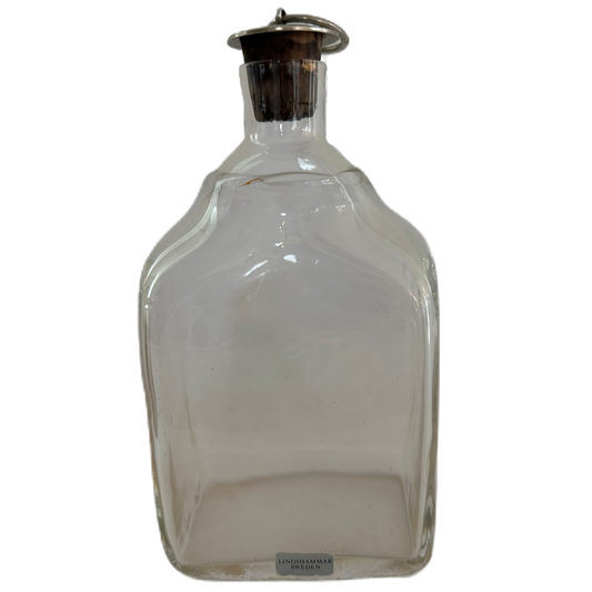 Etikettmärkt plunta i transparent glas med propp i kork och metall från Lindshammar Sweden.